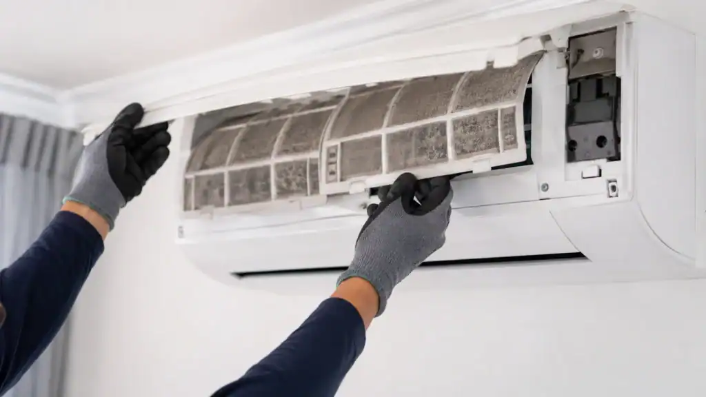 A person wearing gloves opens the front panel of a wall-mounted air conditioner and removes the air filter for cleaning or maintenance.